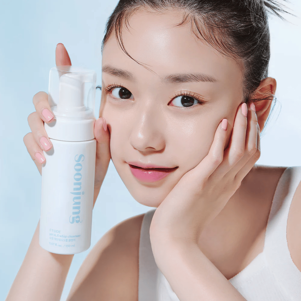 Uma jovem com pele clara segura um frasco de bomba branco com o texto "Soon Jung pH 6.5 Whip Cleanser" de ETUDE. Ela sorri para a câmera com uma maquiagem leve e uma aparência fresca contra um fundo azul claro.