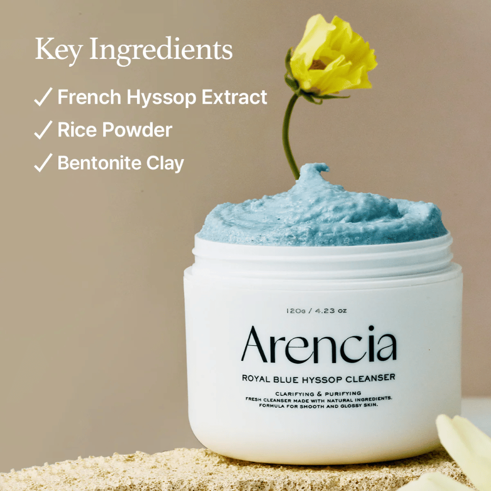 Imagem do frasco "Arencia Royal Blue Hyssop Cleanser" cheio de creme de limpeza azul. Uma flor amarela emerge do creme. O texto na imagem destaca três ingredientes chave: Extrato de Hissopo Francês, Pó de Arroz e Argila Bentonita.