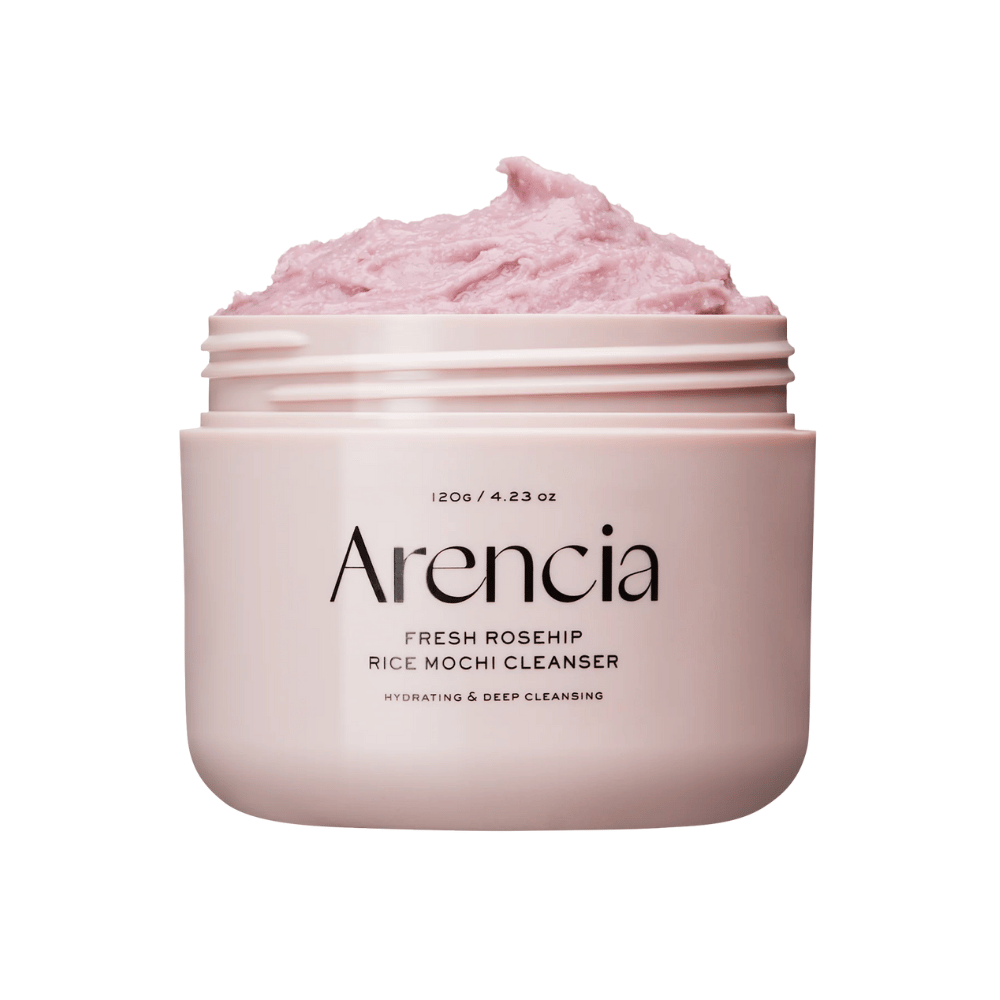 Close-up de um frasco rosa com Arencia Royal Rosehip Cleanser, cheio de creme de limpeza rosa com textura batida, semelhante a mochi. Texto no frasco indica que o produto hidrata e limpa profundamente.