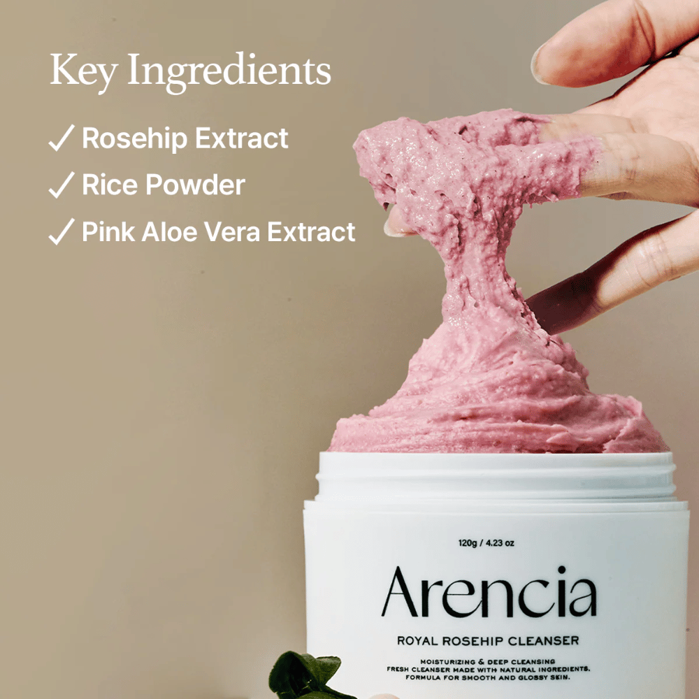 Uma mão levanta a textura de limpeza rosa e fofa do frasco Arencia Royal Rosehip Cleanser, com a lista de ingredientes à esquerda: extrato de rosa mosqueta, pó de arroz e extrato de aloe vera rosa.
