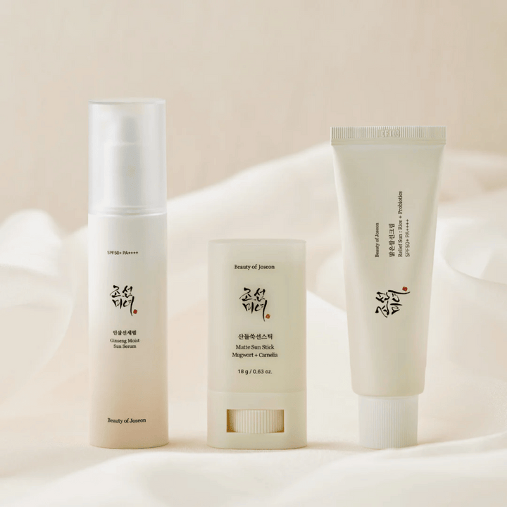 Tre solskyddsprodukter från Beauty of Joseon – Ginseng Moist Sun Serum, Matte Sun Stick och Relief Sun Rice + Probiotics – står uppradade på ett krämfärgat tyg med mjukt ljus.