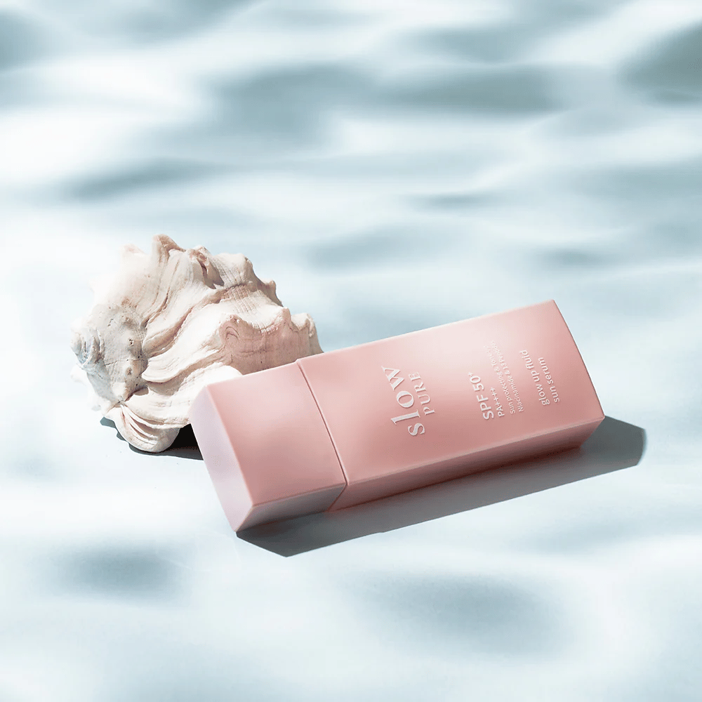 Slow Pure Glow Up Fluid Sun Serum junto a conchas e efeito de água, destaca proteção solar e luminosidade.