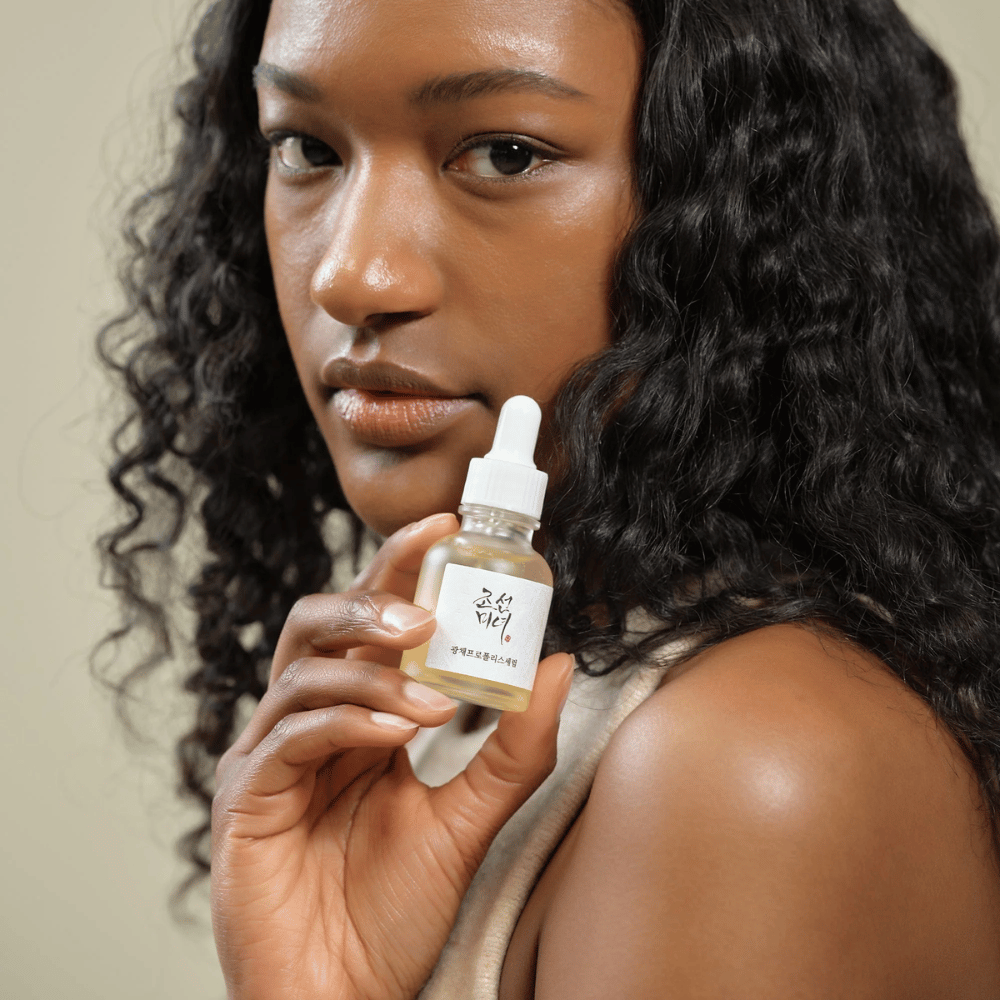 Mulher com cabelo encaracolado segurando um frasco Beauty of Joseon Glow Serum com pipeta, um produto de cuidados com a pele hidratante e iluminador.