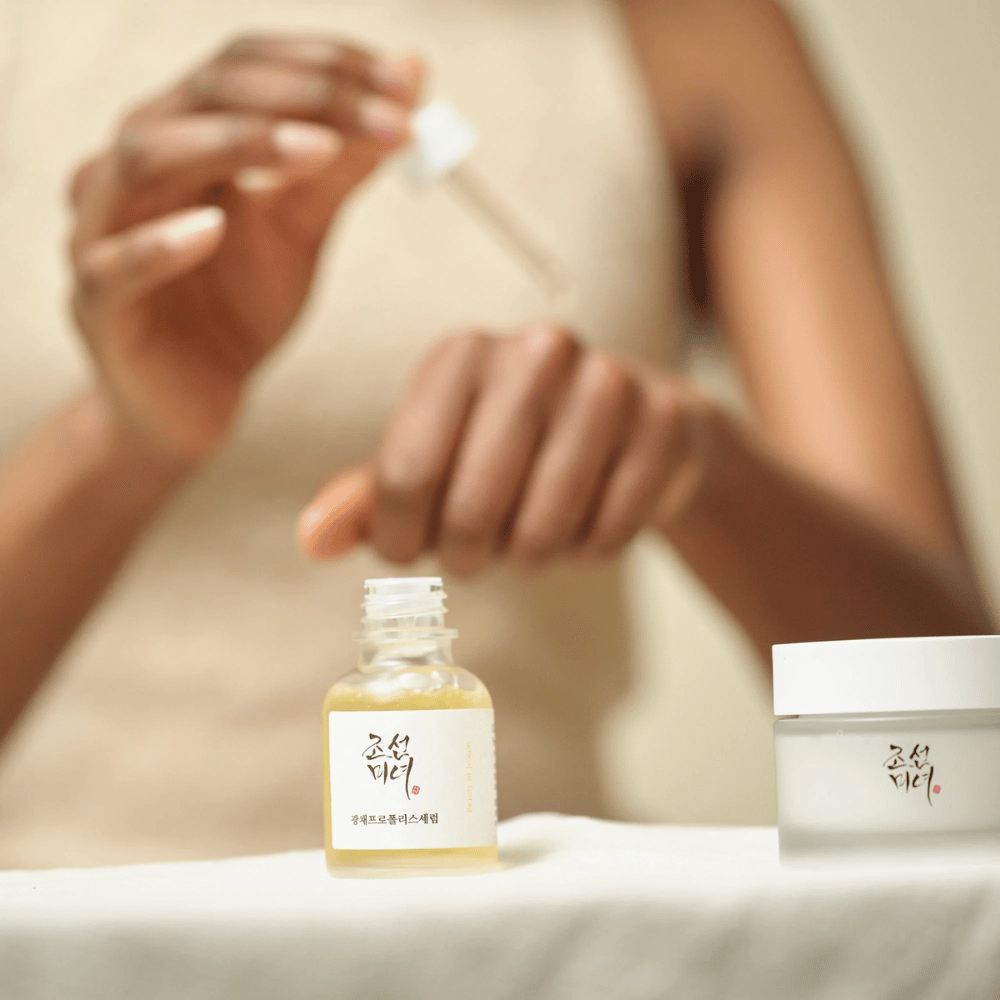 Pessoa aplicando Beauty of Joseon Glow Serum com pipeta de frasco de vidro, com um pote da mesma linha ao lado na mesa.