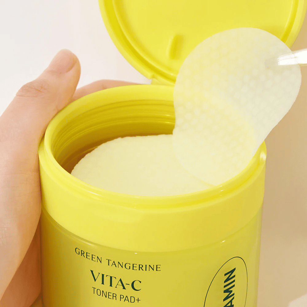 Uma mão usa uma pinça de plástico para pegar um disco de toner texturizado de um recipiente amarelo claro marcado "GREEN TANGERINE VITA-C TONER PAD+" da marca "gooda".