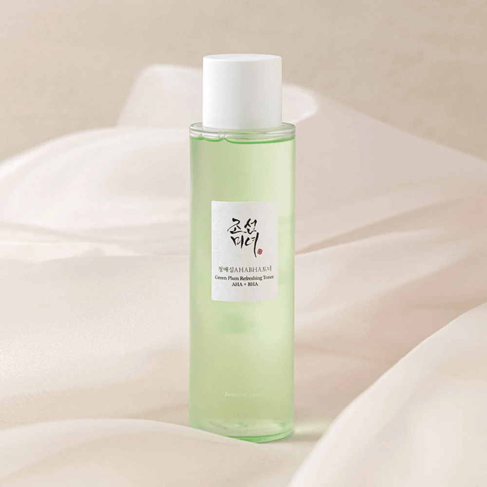 Uma garrafa do "Green Plum Refreshing Toner" AHA + BHA está sobre um fundo suave e claro. O produto tem um líquido verde que reflete frescor e naturalidade. O rótulo é branco com detalhes em preto e vermelho, o que dá um visual elegante e limpo.