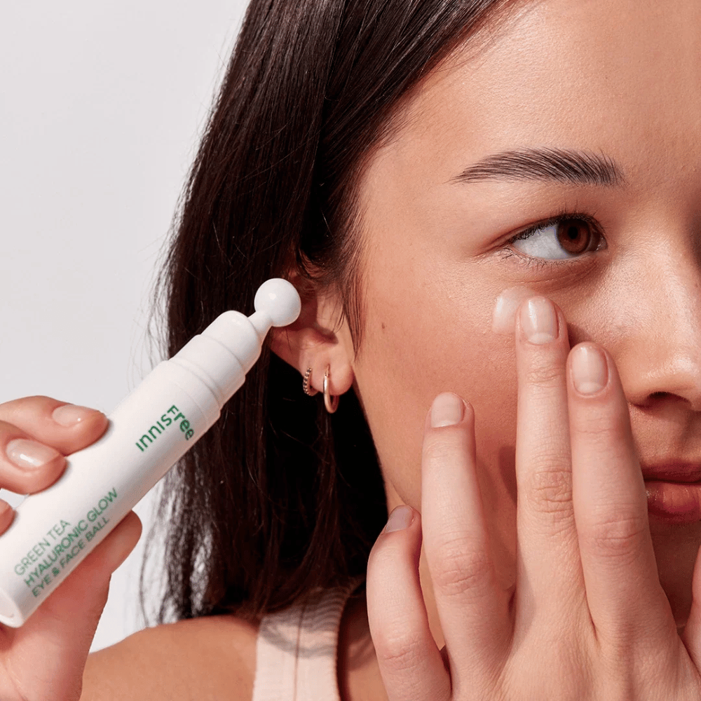 Uma pessoa aplica Innisfree Green Tea Hyaluronic Glow Eye & Face Ball na pele sob os olhos. O produto tem uma parte aplicadora redonda e branca e está em uma garrafa branca com texto verde. O rosto do usuário é parcialmente visível e foca na aplicação do produto.