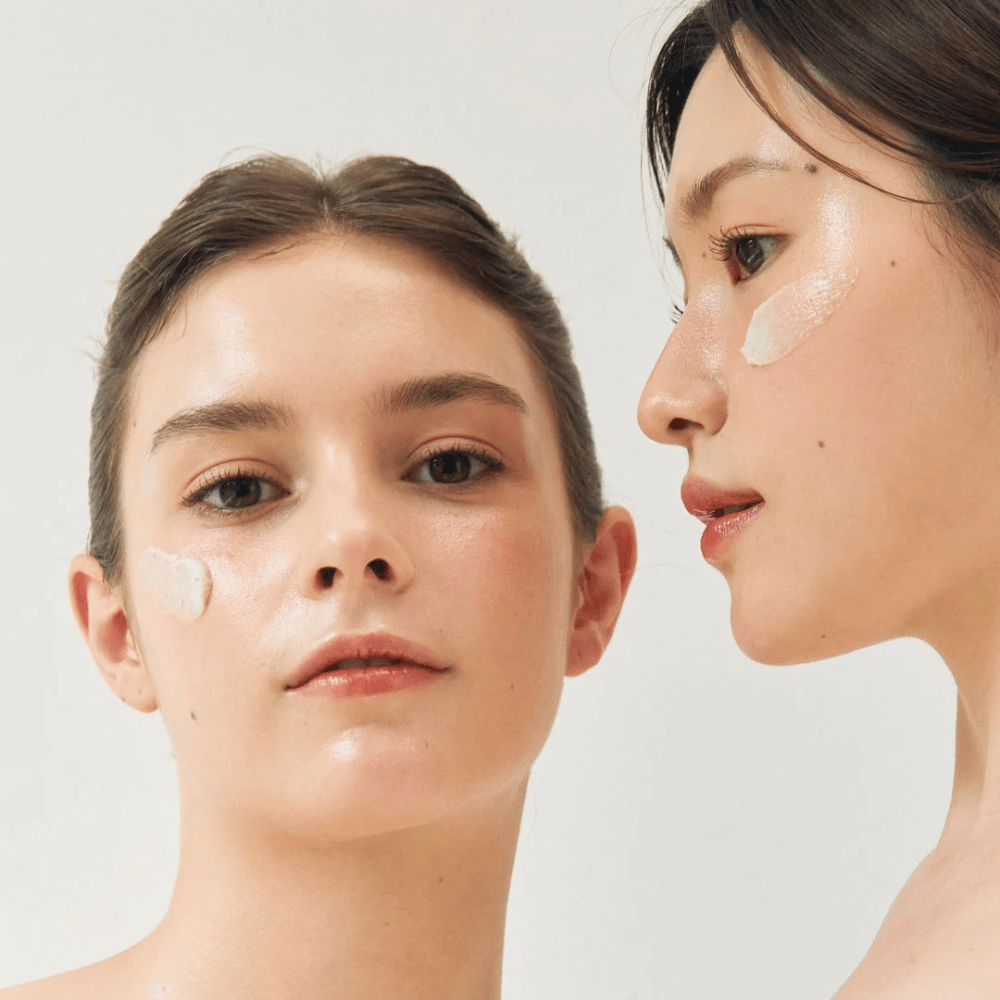 Duas mulheres com máscaras faciais aplicadas em partes de seus rostos. Uma das mulheres tem a máscara na bochecha e no nariz, enquanto a outra a tem debaixo do olho e na bochecha. Elas têm maquiagem natural e parecem frescas, destacando o efeito hidratante e iluminador das máscaras.