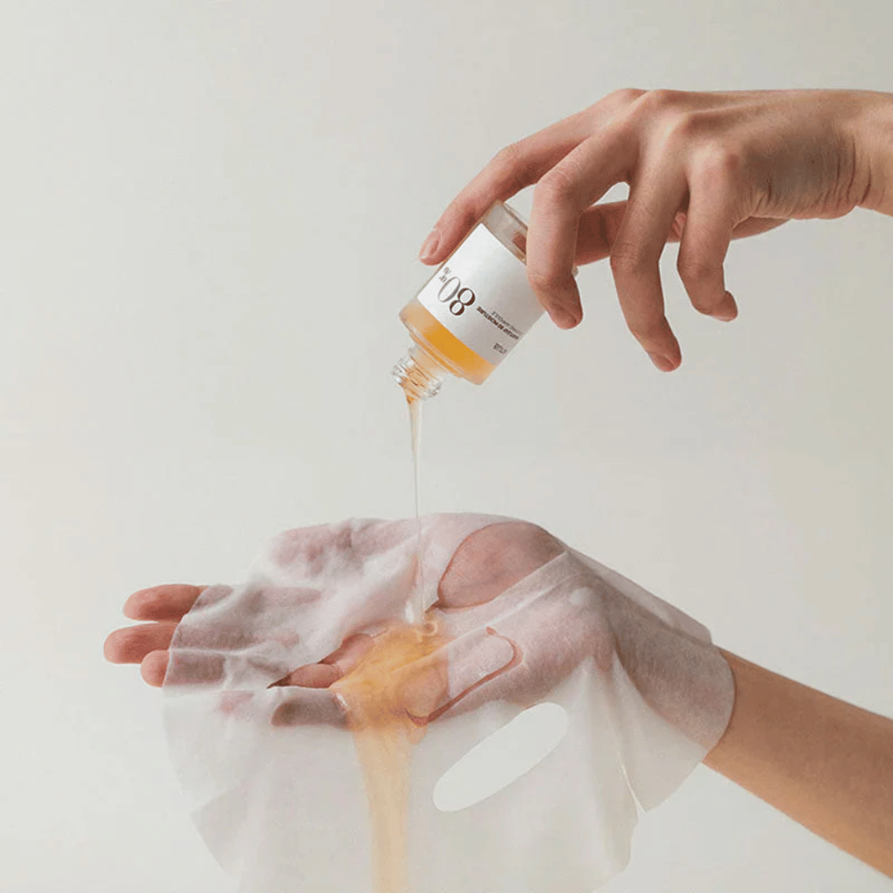 Uma mão segura um líquido dourado e espesso de um pequeno frasco marcado '108' sobre uma máscara facial transparente que é segurada por outra mão. O fundo é neutro e uniforme para destacar o produto.