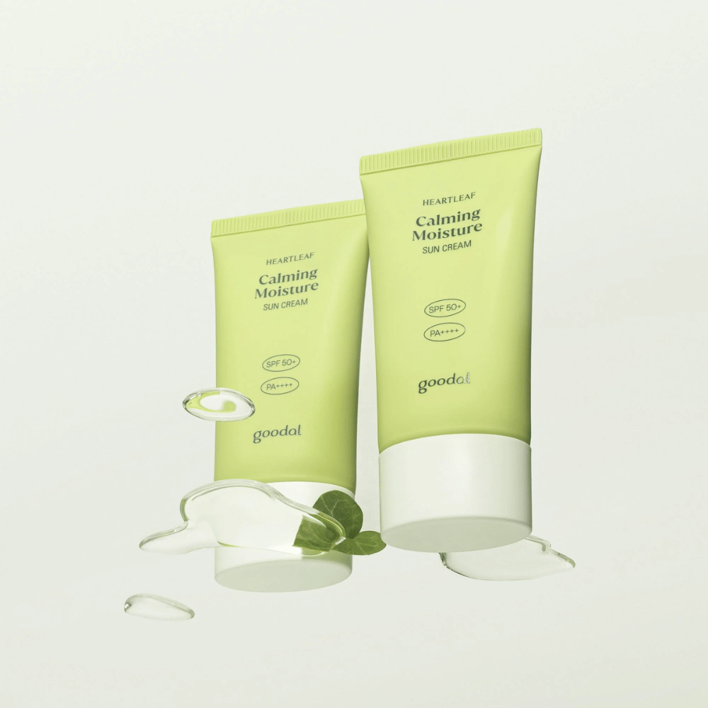 Dois tubos de Goodal Heartleaf Calming Moisture Sun Cream SPF50+ PA++++ rodeados por folhas de plantas e gotas de soro em um fundo claro.