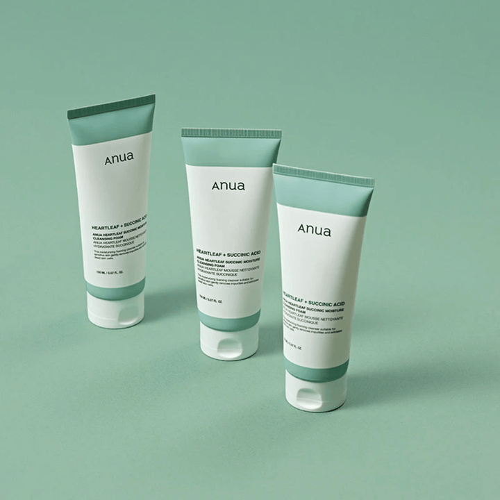 Três embalagens de Anua Heartleaf + Succinic Acid Moisture Cleansing Foam apresentadas sobre um fundo verde-claro. A imagem destaca o design limpo e minimalista do produto, além de transmitir uma impressão profissional da gama de produtos.