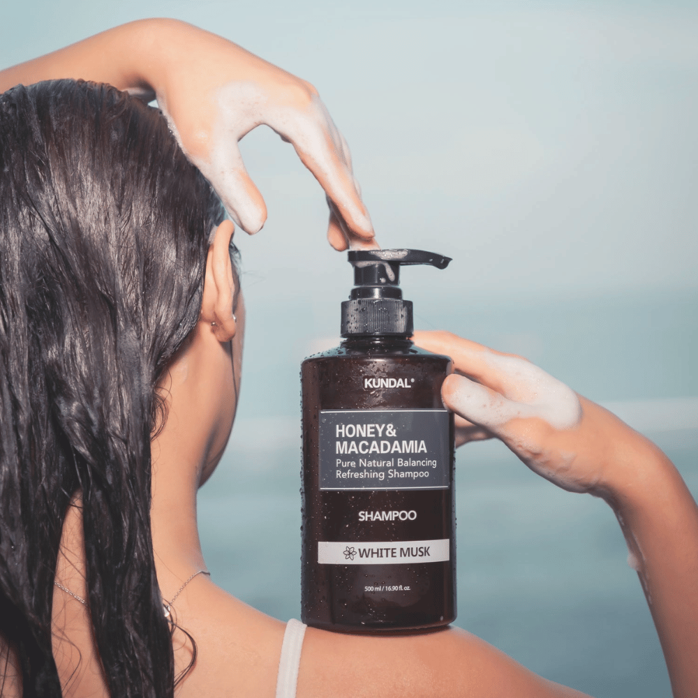 Mulher com cabelo molhado aplica espuma e segura KUNDAL Honey & Macadamia Nature Shampoo White Musk com fundo do mar.