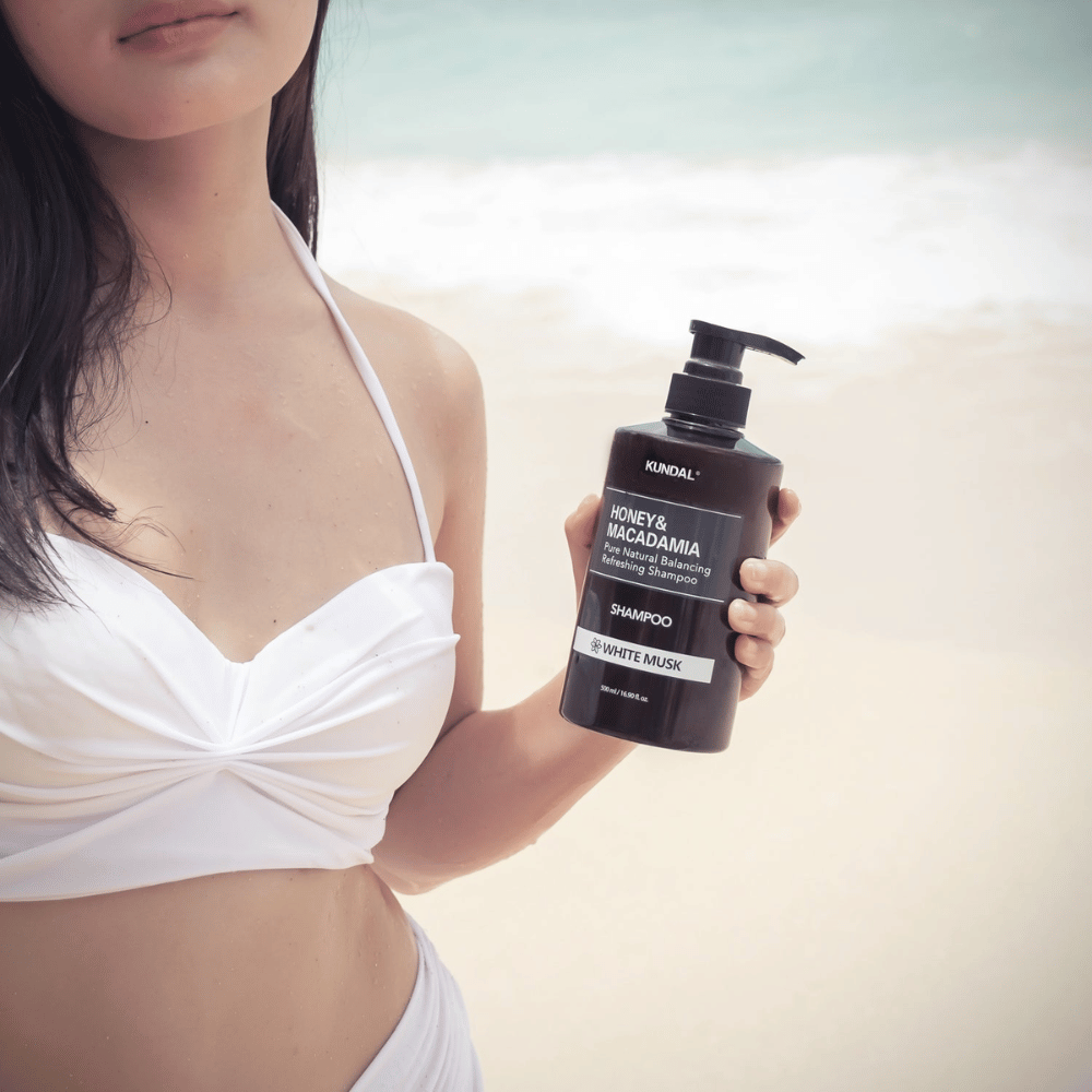 Mulher de biquíni branco segura KUNDAL Honey & Macadamia Nature Shampoo White Musk numa praia com o mar ao fundo.