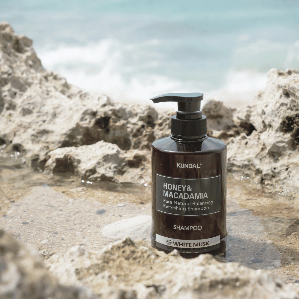 KUNDAL Honey & Macadamia Nature Shampoo White Musk colocado sobre rochas junto ao mar, um produto de cuidado capilar natural com fragrância fresca.
