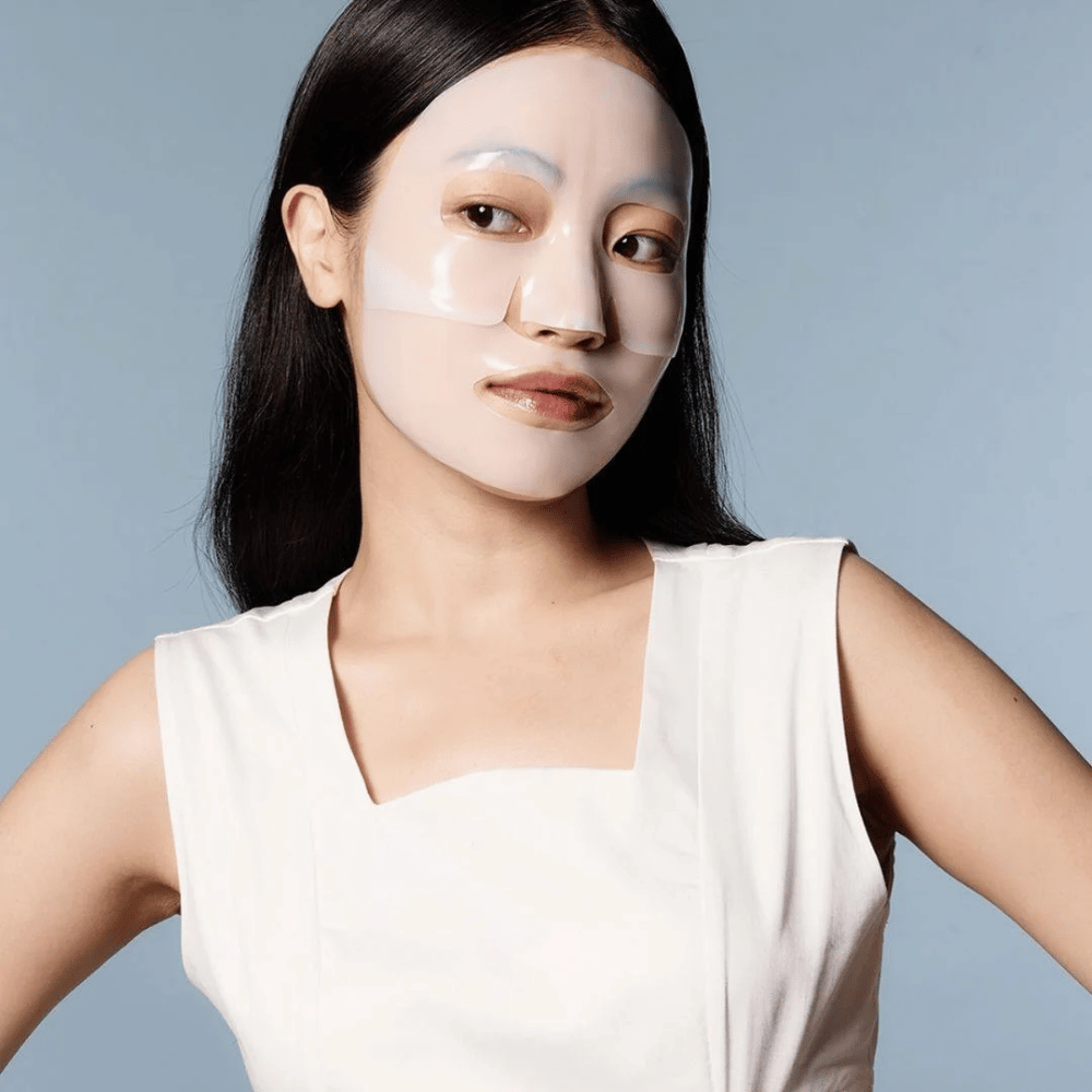 Uma mulher em um vestido branco sem mangas usa uma máscara facial transparente que cobre quase todo o seu rosto. A máscara dá uma impressão de cuidados avançados com a pele e oferece a aparência de um tratamento de beleza, projetada para hidratar profundamente e tratar a pele.