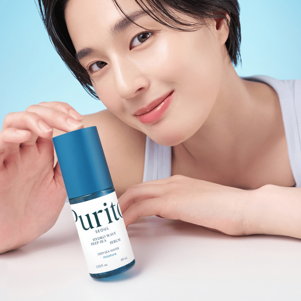 Uma mulher com cabelo curto segura um soro azul de Purito, Hydro Wave Deep Sea Serum. Sua pele parece radiante e úmida, destacando o produto em foco. O fundo é azul claro, proporcionando uma sensação fresca e limpa. O produto é aprofundado no cuidado da pele.