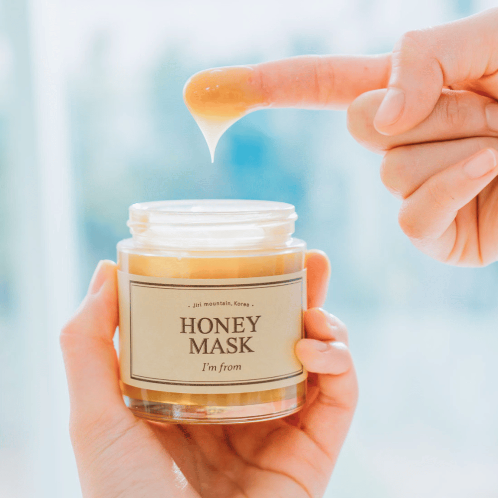 Uma mão segura um frasco aberto de "I'm from Honey Mask", e um dedo mergulha na máscara dourada contra um fundo claro.