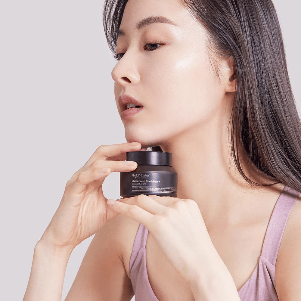 Uma mulher segura um frasco de MARY & MAY Idebenone Blackberry Intense Cream. Ela tem uma pele radiante e sorri levemente, vestida com um top sem mangas. O produto é um creme intenso com idebenona e complexo de amora destinado à firmeza e hidratação da pele.