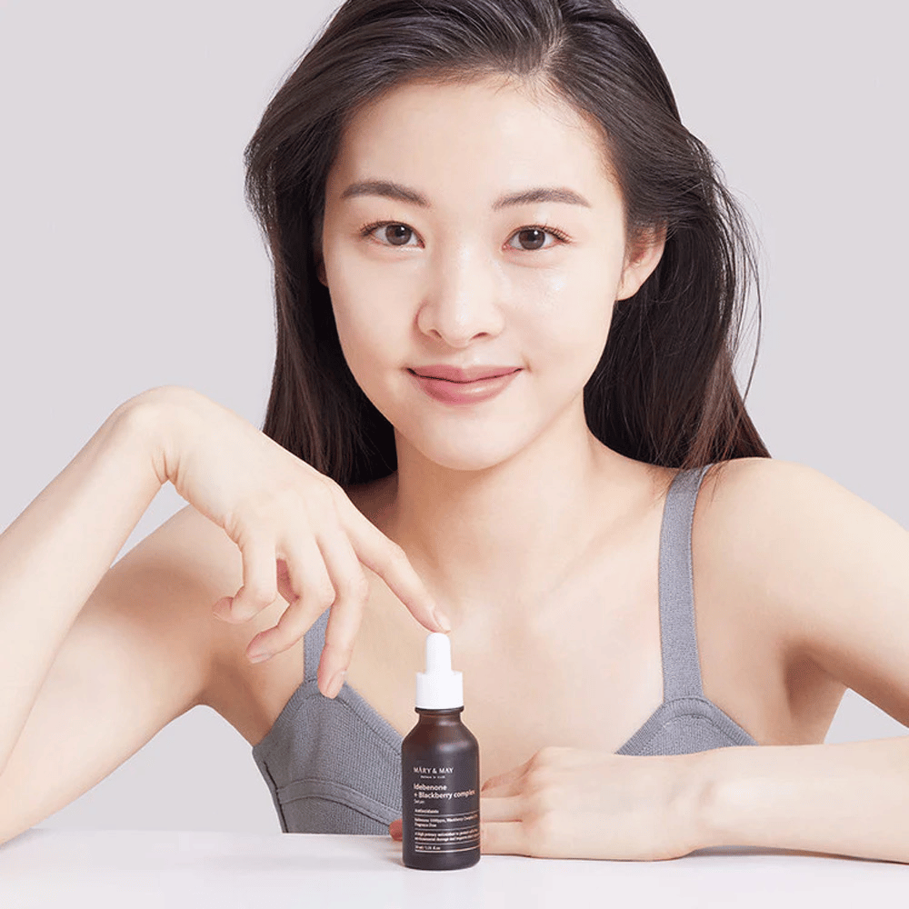 Uma mulher com cabelo comprido e escuro segura o MARY & MAY Idebenone + Blackberry Complex Serum, uma garrafa de vidro escuro com conta-gotas. Ela tem um sorriso alegre e aponta para a garrafa com a outra mão. A mulher está vestida com um top cinza sem mangas e parece estar em um ambiente claro e simples, destacando o foco do produto em cuidados com a pele e beleza.