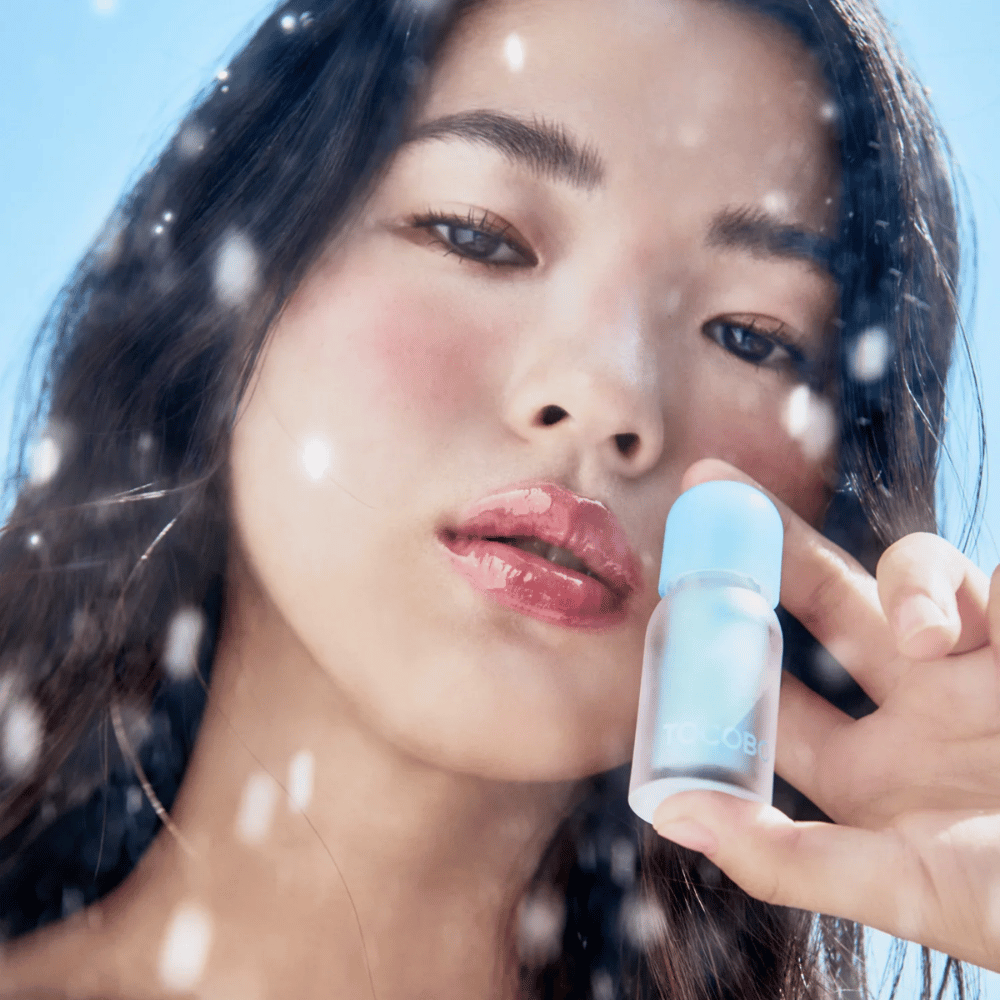 Kvinna håller TOCOBO Juicy Berry Plumping Lip Oil i handen under klar himmel. Läppoljan ger glans, fukt och en kylande, fräsch effekt.