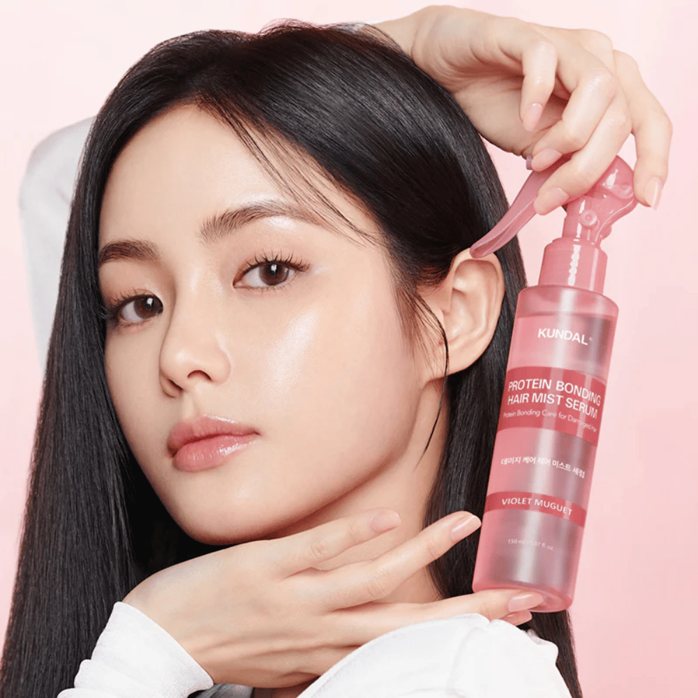 Mulher com cabelo preto longo segurando KUNDAL Protein Bonding Hair Mist Serum Violet Muguet em frente ao rosto, contra um fundo rosa claro.