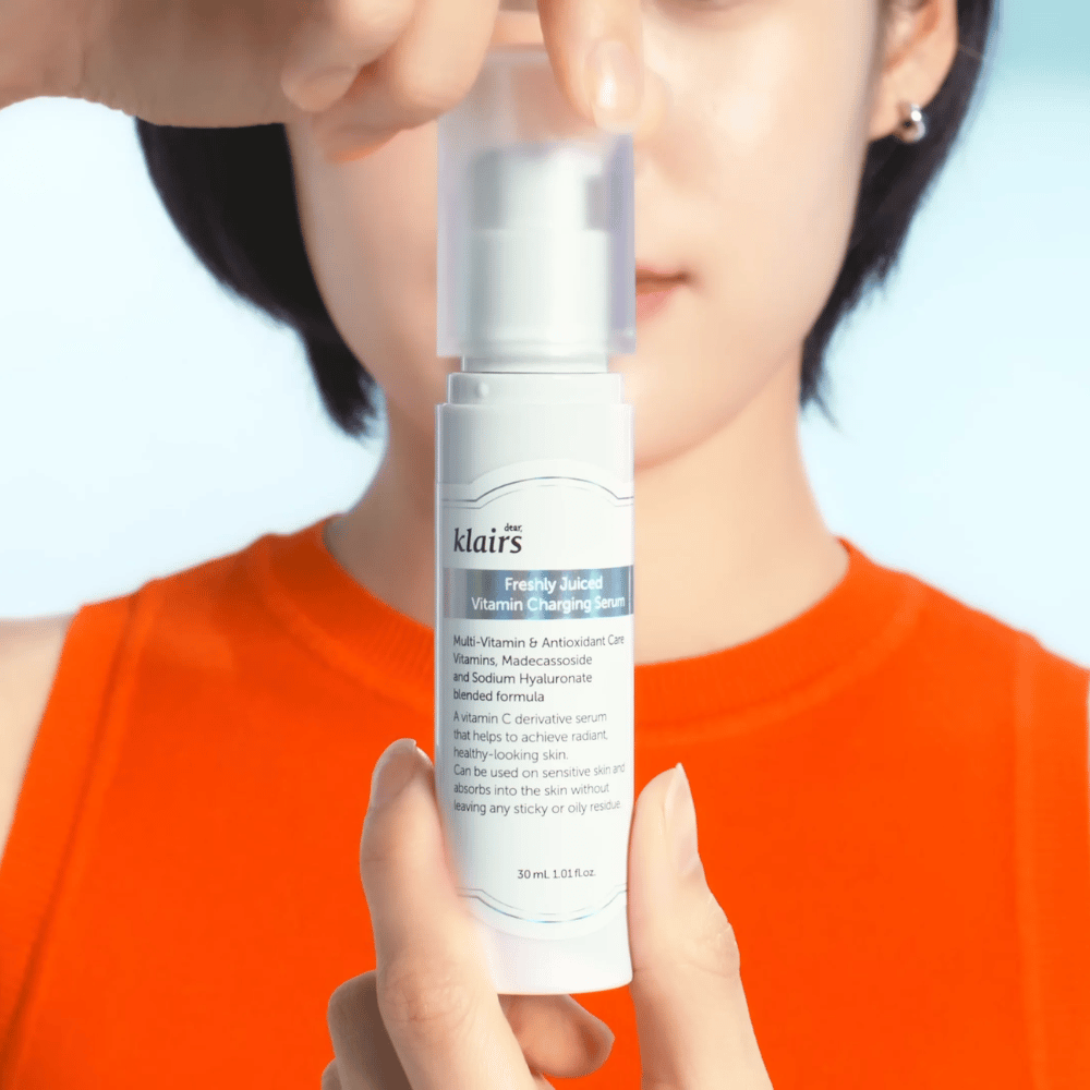 Mulher com camisa laranja segura Klairs Freshly Juiced Vitamin Charging Serum à sua frente contra um fundo azul.