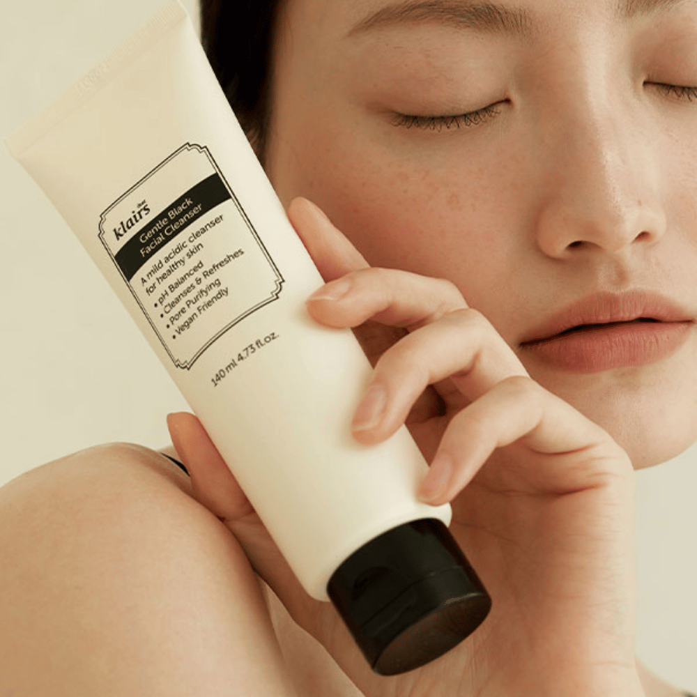 Uma pessoa segura um tubo de Klairs Gentle Black Facial Cleanser contra sua bochecha, olhos fechados e tom de pele natural visíveis ao fundo.