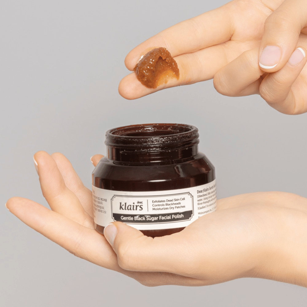 Mãos segurando um frasco aberto de Klairs Gentle Black Sugar Facial Polish e mostrando o produto em um dedo, sobre um fundo cinza.