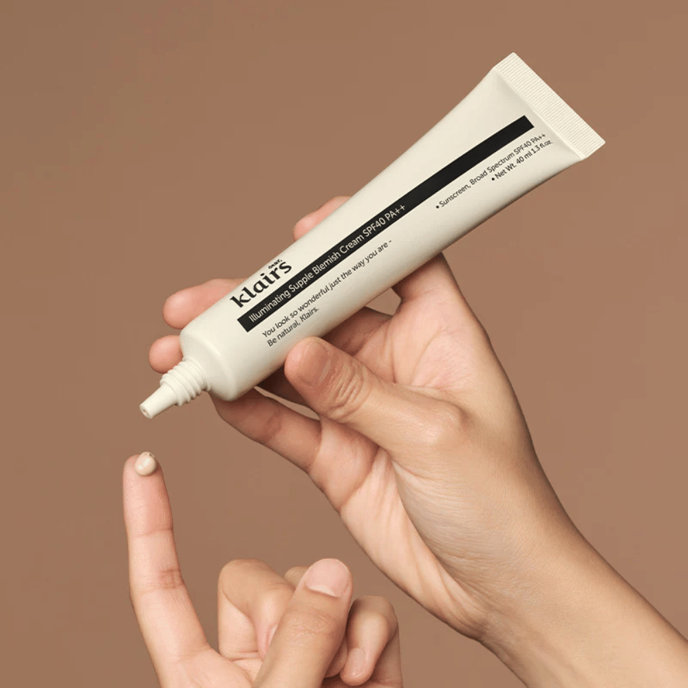 Uma mão segura um tubo de Klairs Illuminating Supple Blemish Cream SPF40 PA++ e espreme uma pequena quantidade de creme num dedo, contra um fundo castanho.