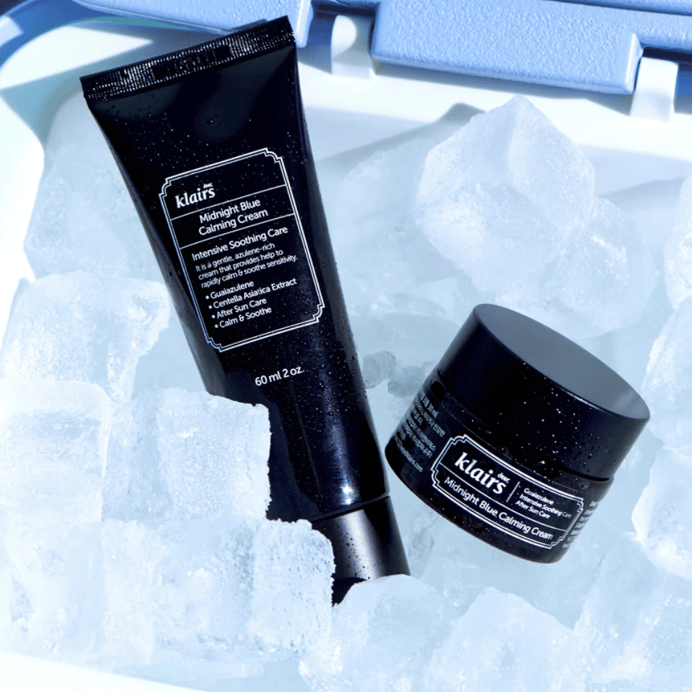 Um tubo e um pote de Klairs Midnight Blue Calming Cream estão sobre cubos de gelo, com foco em propriedades calmantes e hidratantes, contra um fundo azul.