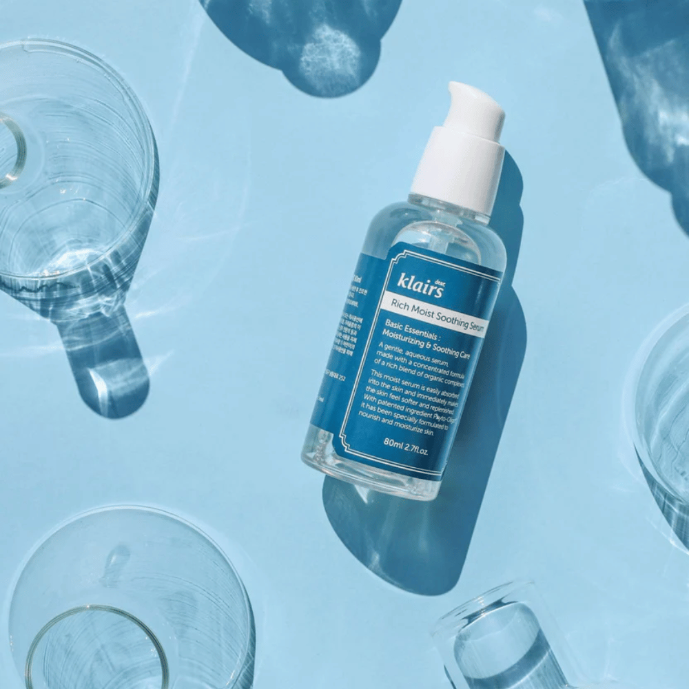 Uma garrafa Klairs Rich Moist Soothing Serum entre copos de água com reflexos sobre um fundo azul.