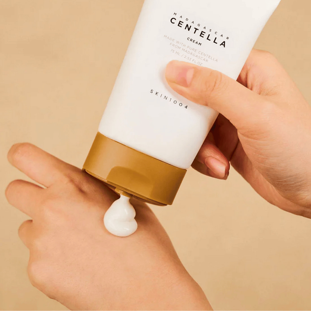 A imagem mostra uma pessoa segurando um tubo de "Madagascar Centella Cream" de SKIN1004. É um creme para a pele feito de Centella pura de Madagascar, no tamanho de 75 ml / 2.53 fl.oz. O creme é aplicado na mão e é destinado a nutrir e hidratar a pele.