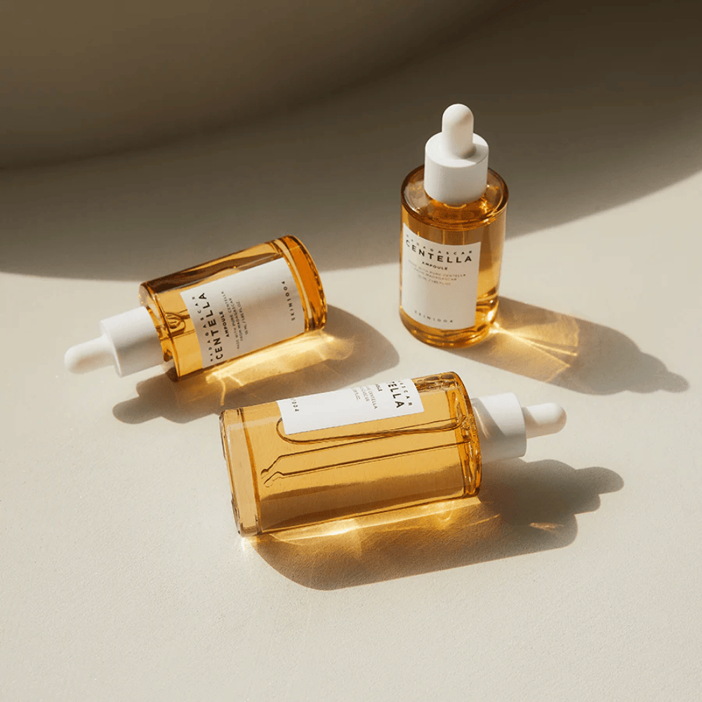 A imagem mostra três garrafas de "Madagascar Centella Asiatica 100 Ampoule". Elas estão colocadas em uma superfície onde a luz do sol cria um belo reflexo através do conteúdo dourado nas garrafas. O produto parece ser parte de uma linha de cuidados com a pele que utiliza centella asiatica, um ingrediente conhecido por suas propriedades calmantes. O design das garrafas é simples e moderno, o que sugere que é uma linha de cuidados com a pele premium.