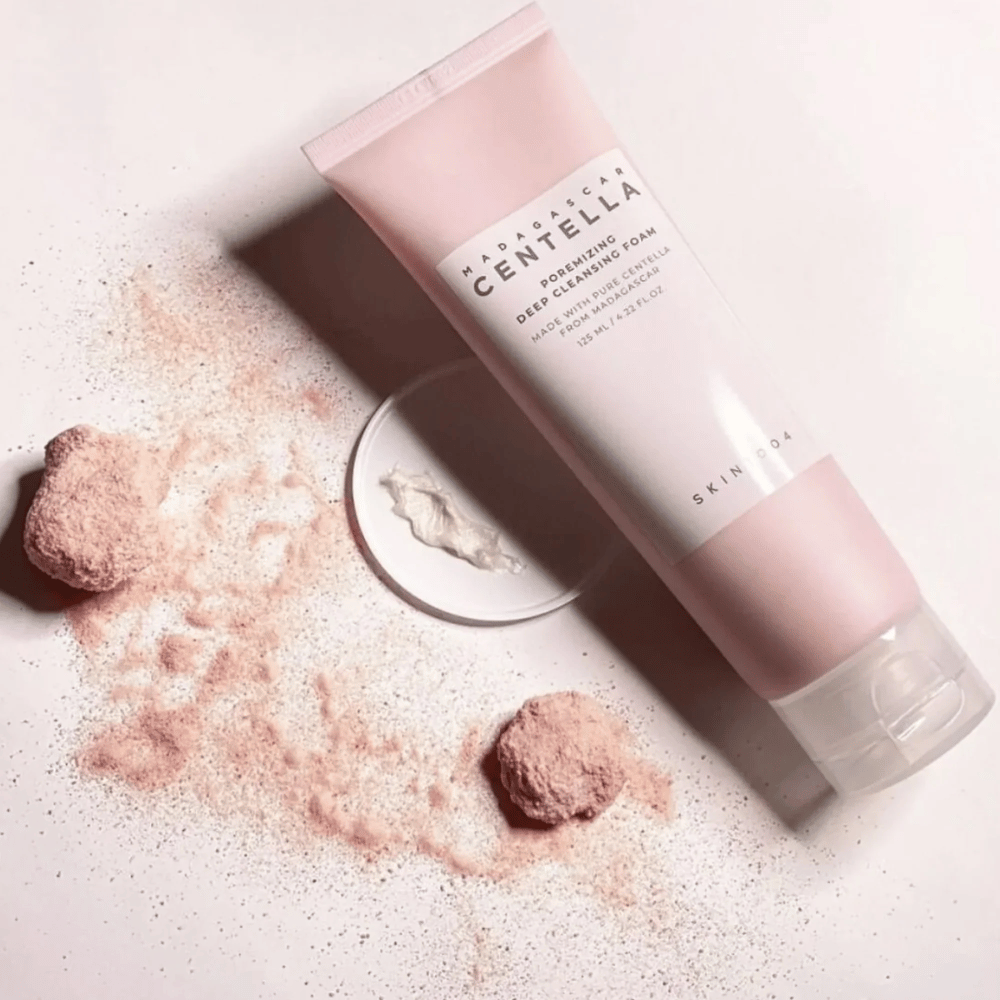 Madagascar Centella Poremizing Deep Cleansing Foam é apresentado em um tubo rosa. Os tubos estão sobre uma superfície clara junto com pó rosa que dá uma sensação de pureza e frescor. O produto é projetado para limpar profundamente a pele com a ajuda de centella pura de Madagascar.