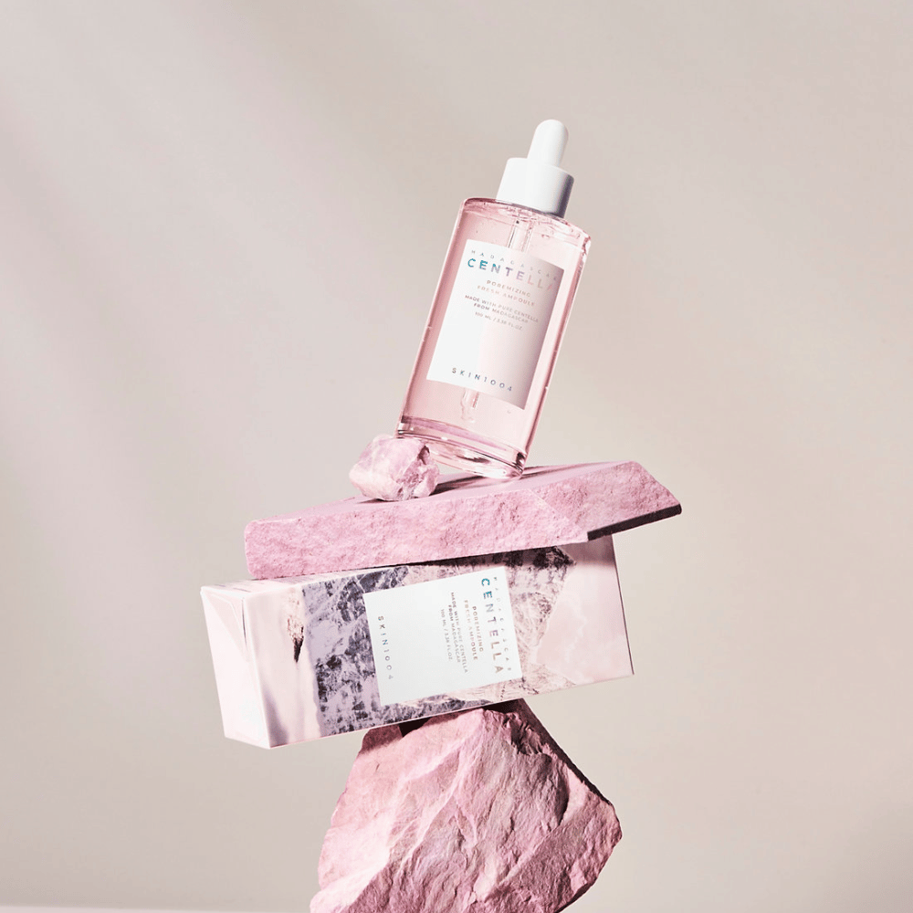Frasco transparente com ampola rosa de Centella da SKIN1004, equilibrado em blocos de pedra, com uma embalagem de papelão embaixo, contra um fundo neutro.