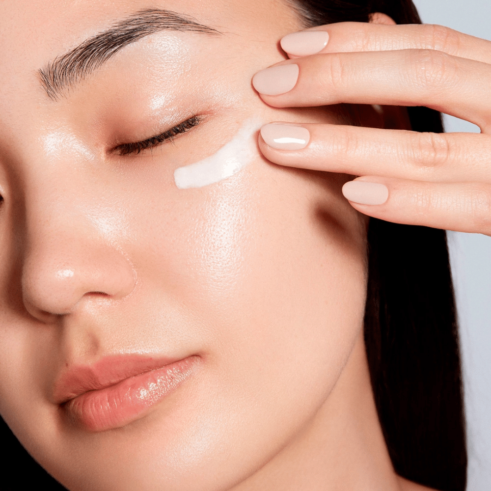 Uma mulher aplica cuidadosamente um creme branco para os olhos sob o olho. Sua pele parece radiante e hidratada, o que enfatiza o efeito do creme para os olhos. Isso destaca o produto como tanto hidratante quanto adequado para a pele sensível ao redor dos olhos.