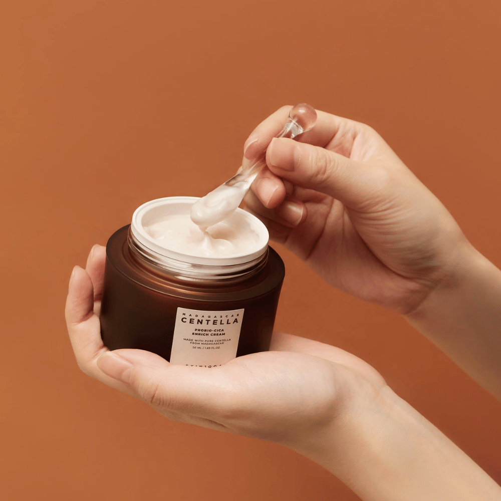 Mãos segurando um frasco aberto de Madagascar Centella Probio-Cica Enrich Cream e uma espátula com creme contra um fundo uniforme de cor terracota.