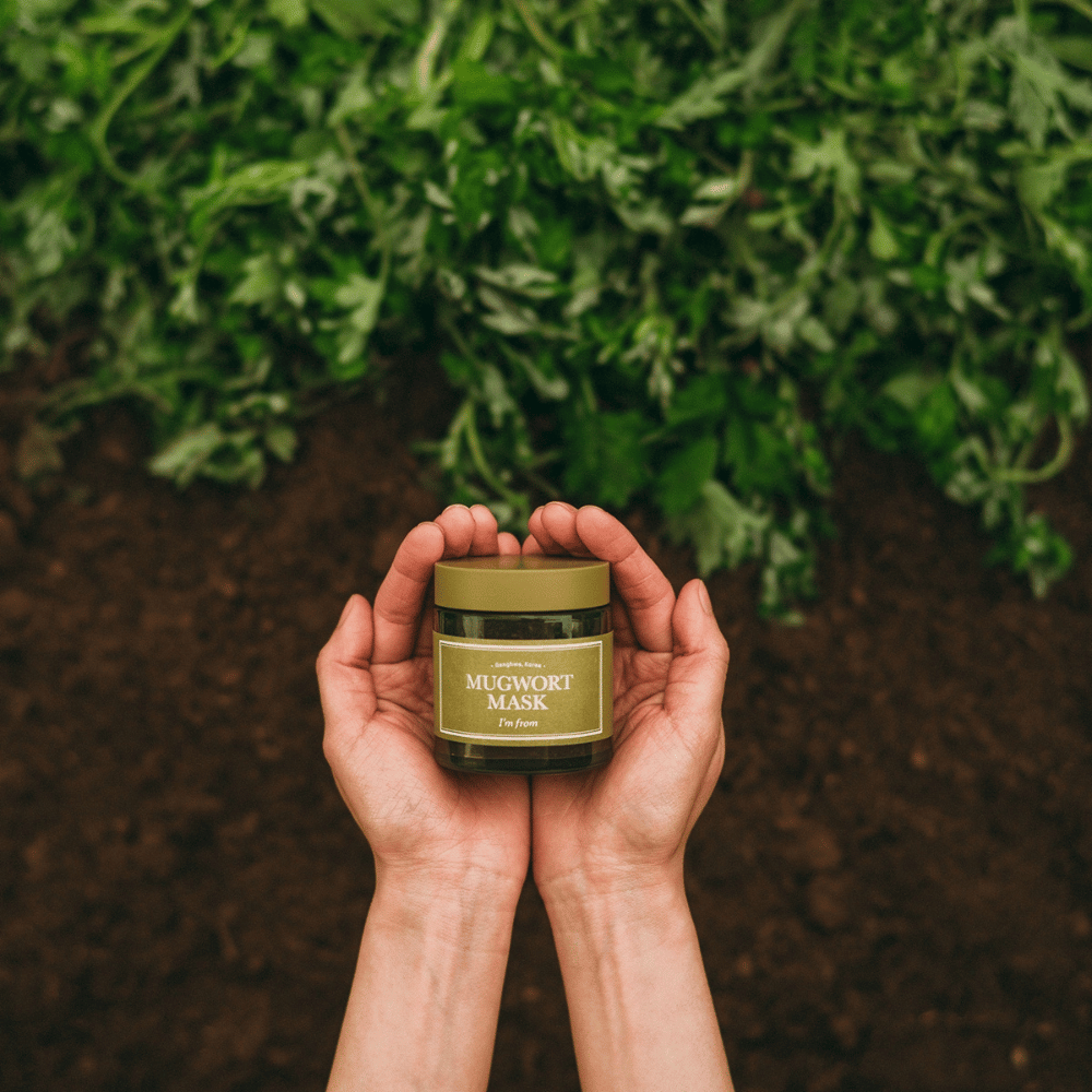 Mãos segurando I'm From Mugwort Mask contra um fundo de terra e ervas frescas, destacando os ingredientes naturais do produto.