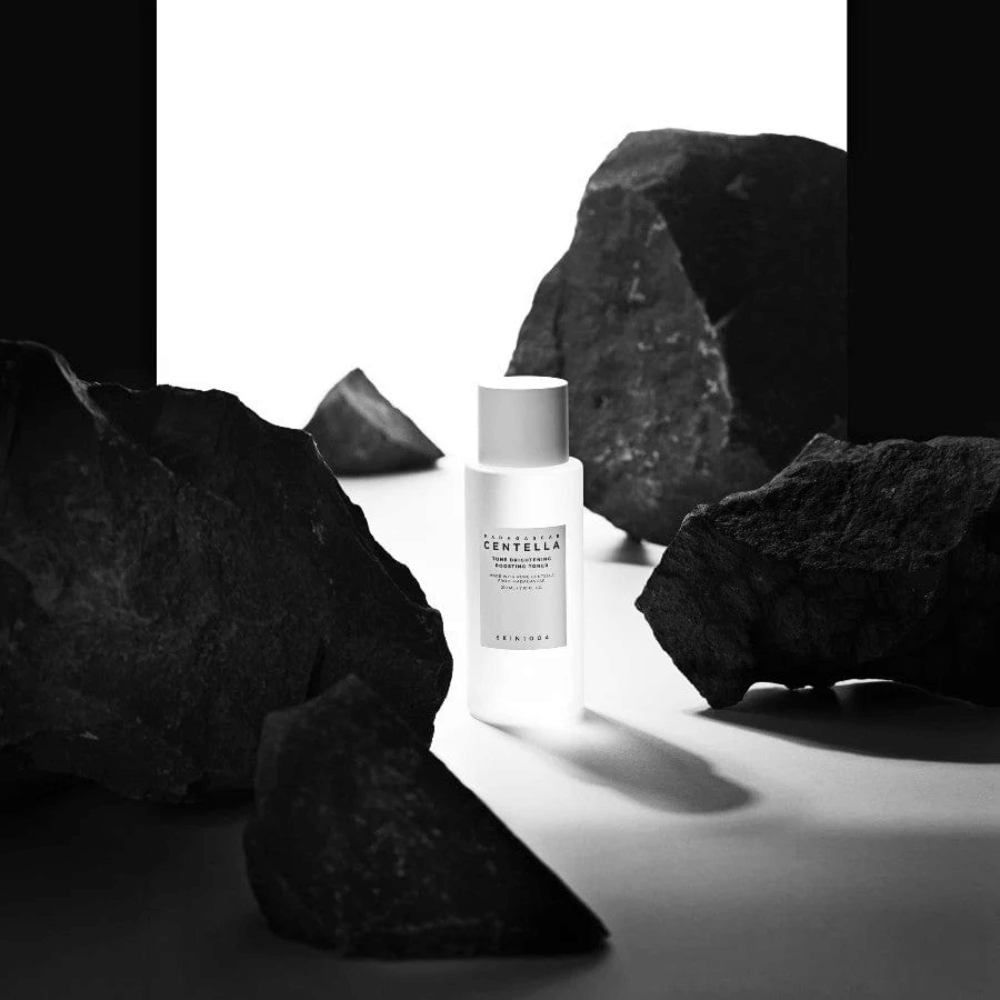 Frasco branco SKIN1004 Madagascar Centella Tone Brightening Boosting Toner  colocado de forma dramática entre grandes pedras pretas contra um fundo branco.