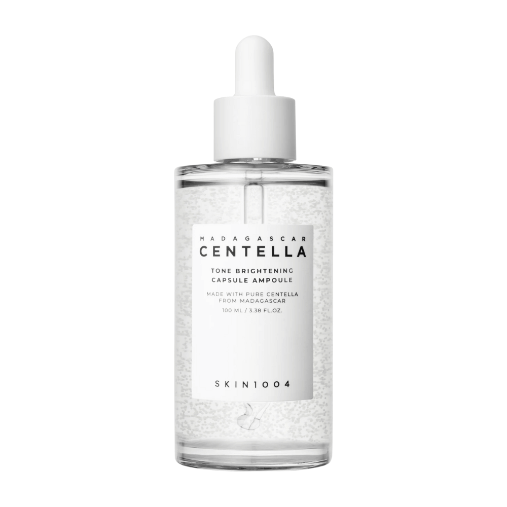 A imagem mostra uma garrafa de "SKIN1004 Madagascar Centella Tone Brightening Capsule Ampoule". É uma garrafa transparente com um aplicador de gotas branco. Na etiqueta, diz que o produto contém ingredientes puros de centella de Madagascar. O conteúdo da garrafa parece ter pequenas cápsulas ou partículas, o que pode ser parte de suas propriedades de cuidados com a pele iluminadoras. A garrafa contém 100 ml / 3,38 fl.oz. do produto.