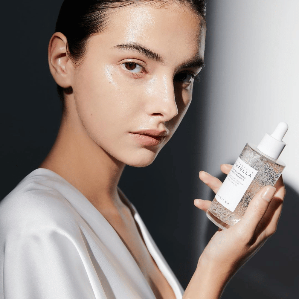 Na imagem, vê-se uma pessoa segurando uma garrafa com aplicador de gotas. A garrafa está marcada "SKIN1004 Madagascar Centella Tone Brightening Capsule Ampoule". A pessoa tem um tom de pele cintilante que reflete a luz, o que sugere um possível efeito do produto.