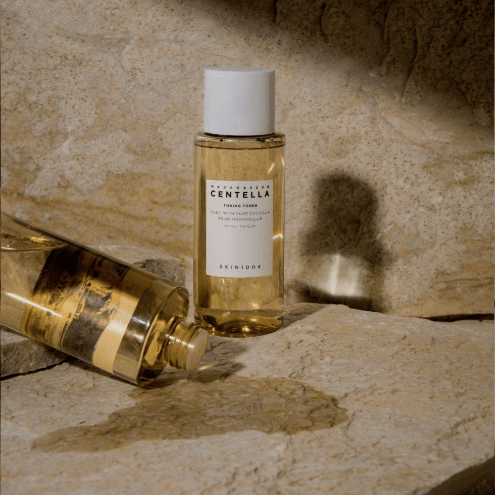 A imagem mostra uma garrafa "Madagascar Centella Toning Toner" de SKIN1004. É uma garrafa transparente cheia de um líquido amarelado, provavelmente um toner para cuidados com a pele. Diz-se que é feito com centella pura de Madagascar e o conteúdo é de 210 ml / 7.10 fl.oz. A garrafa e uma garrafa deitada estão colocadas sobre uma superfície rochosa, o que dá uma sensação natural e minimalista.