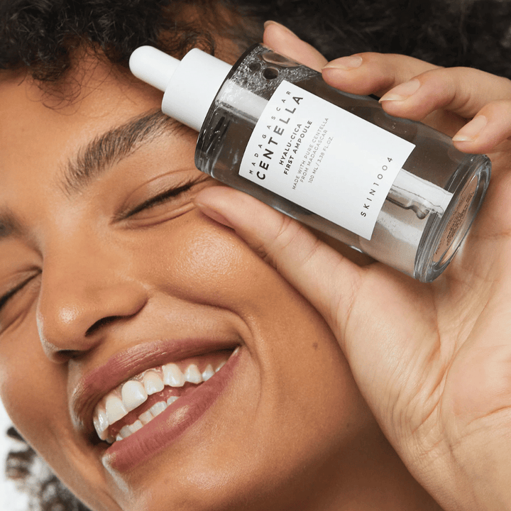 Leende kvinna håller upp Skin1004 Madagascar Centella Hyalu-Cica First Ampoule bredvid ansiktet.