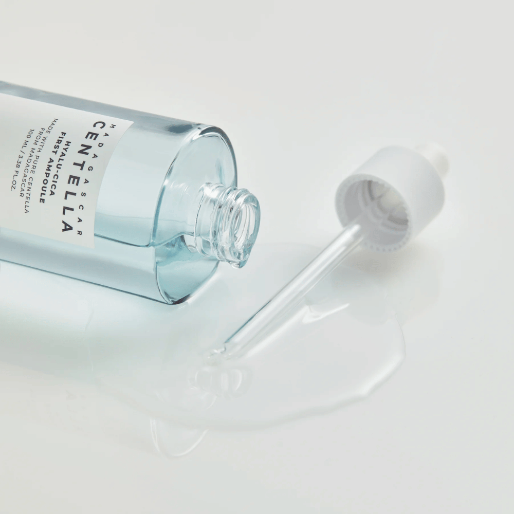 Close-up de frasco aberto Skin1004 Madagascar Centella Hyalu-Cica First Ampoule com pipeta deitada e soro espalhado sobre superfície branca.