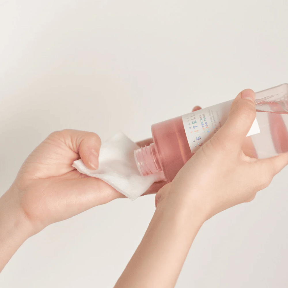 Uma pessoa despeja Madagascar Centella Poremizing Clear Toner de uma garrafa transparente em um disco de algodão. O produto, que tem um tom rosa, é usado para refrescar e limpar a pele. As mãos são visíveis, mostrando a aplicação do toner.