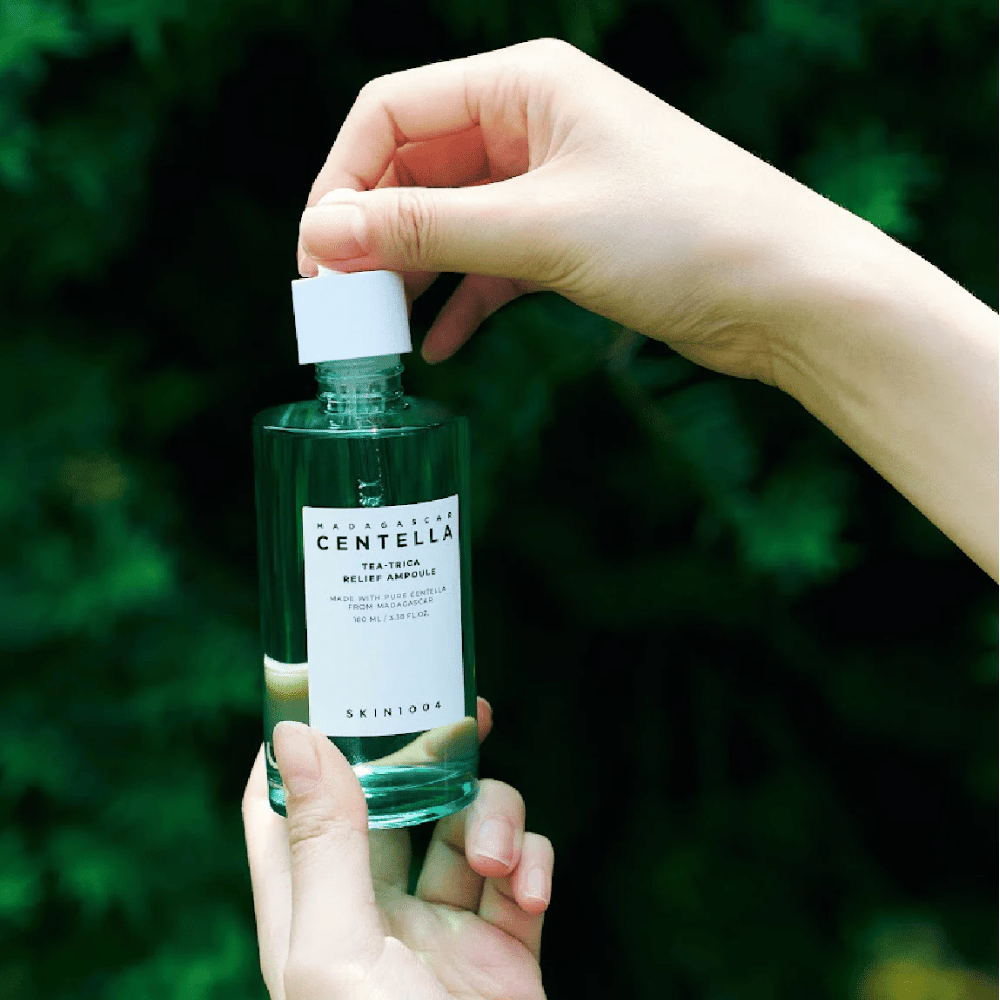 Pessoa abrindo SKIN1004 Madagascar Centella Tea-Trica Relief Ampoule – garrafa de vidro verde com rótulo branco segurada nas mãos em frente a um fundo verdejante.