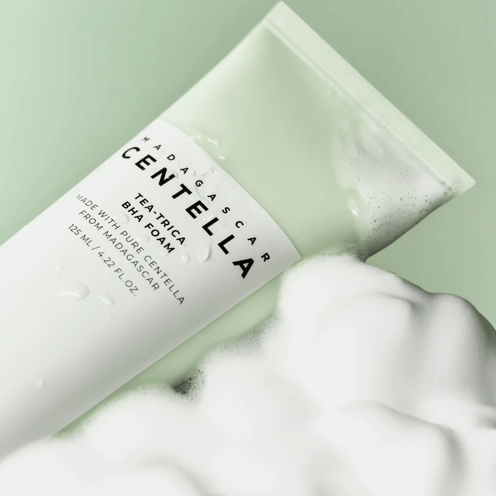 Um produto do tipo espuma de limpeza, "Madagascar Centella Tea-trica BHA Foam", é parcialmente visível com espuma ao redor. O produto tem um design verde e branco, e o texto "Madagascar Centella" e "Tea-trica BHA Foam" são claramente visíveis. O volume é de 125 ml.