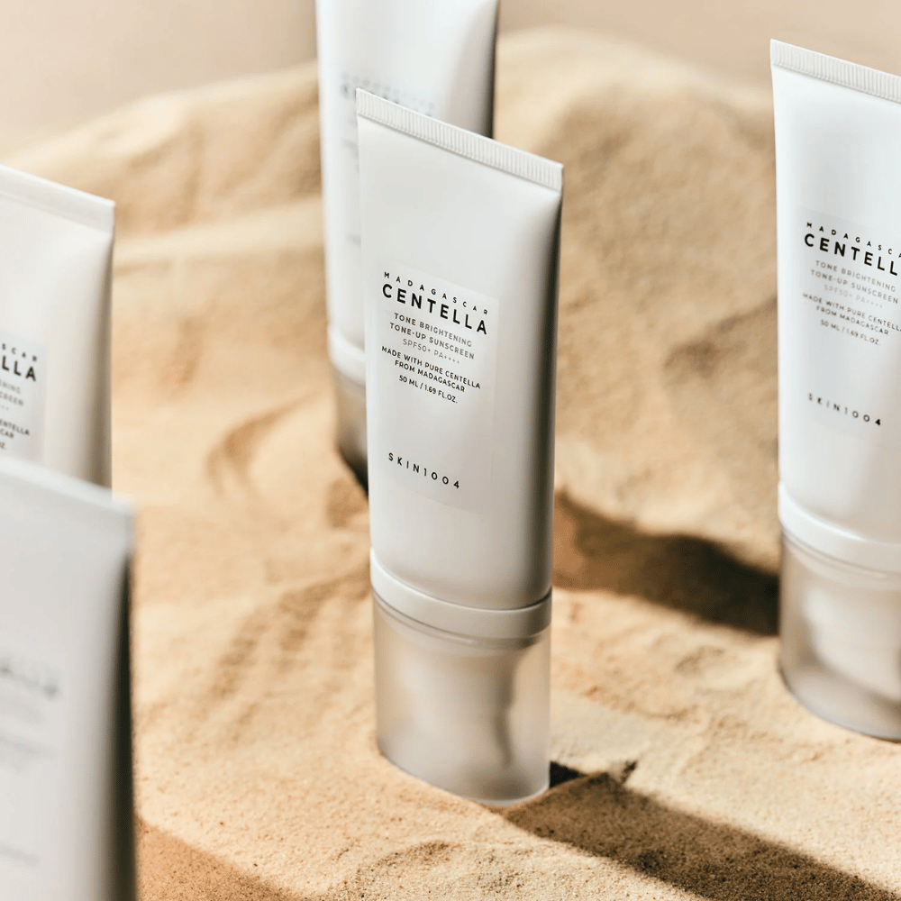 Vários tubos de Madagascar Centella Tone Brightening Tone-Up Sunscreen estão na areia. O tubo é branco com texto preto e contém 50 ml. O produto tem SPF 50+ PA++++ e é fabricado com Centella pura de Madagascar.