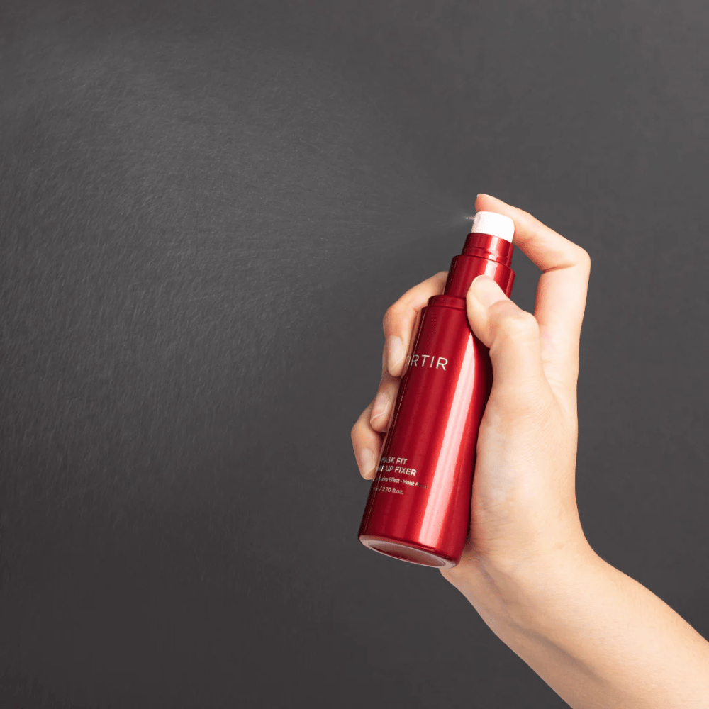 Uma mão segurando uma garrafa vermelha da marca tirtir Mask Fit Make Up Fixer e pulverizando uma névoa fina, ideal para fixar maquiagem.