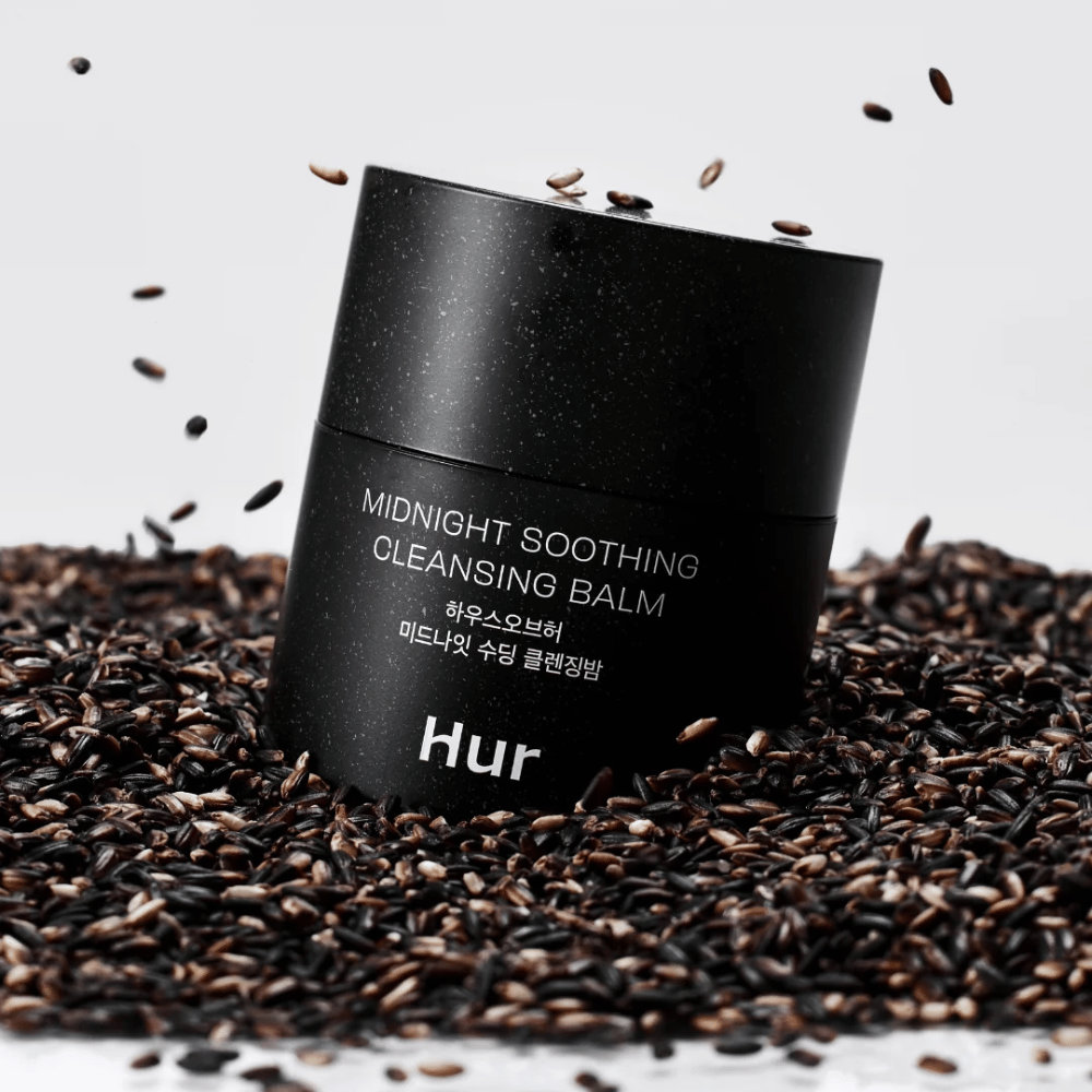 Como Midnight Soothing Cleansing Balm em pote preto colocado sobre sementes pretas. Bálsamo de limpeza calmante que dissolve maquiagem e impurezas enquanto cuida da pele.